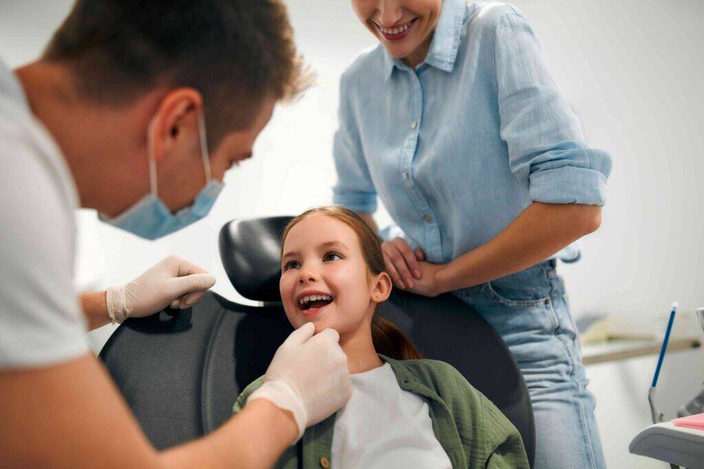 Dentist examining a smiling child’s teeth while a parent looks on.